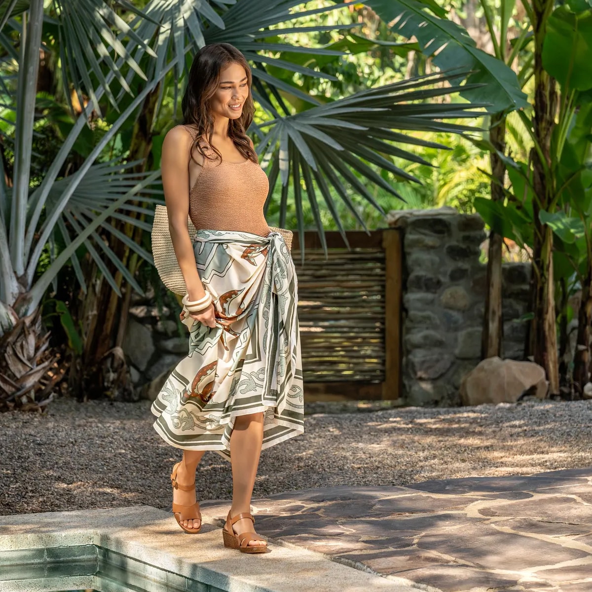 Woman walking alongside a pool wearing the Nancy low wedge sandal in brown.