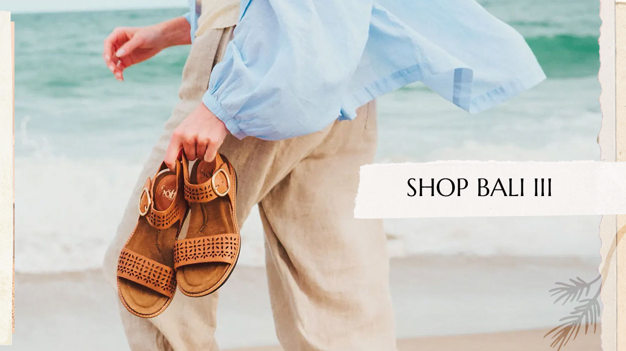 Woman wearing a blue shirt and white pants holding a pair of the Bali II sandal in brown. Shop the Bali III.