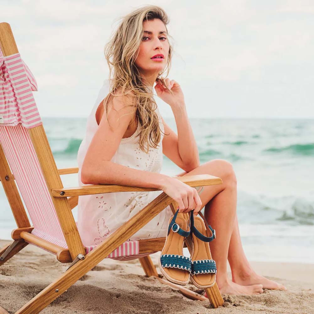 Model sitting on the beach holding a pair of the Inez sandal in blue.