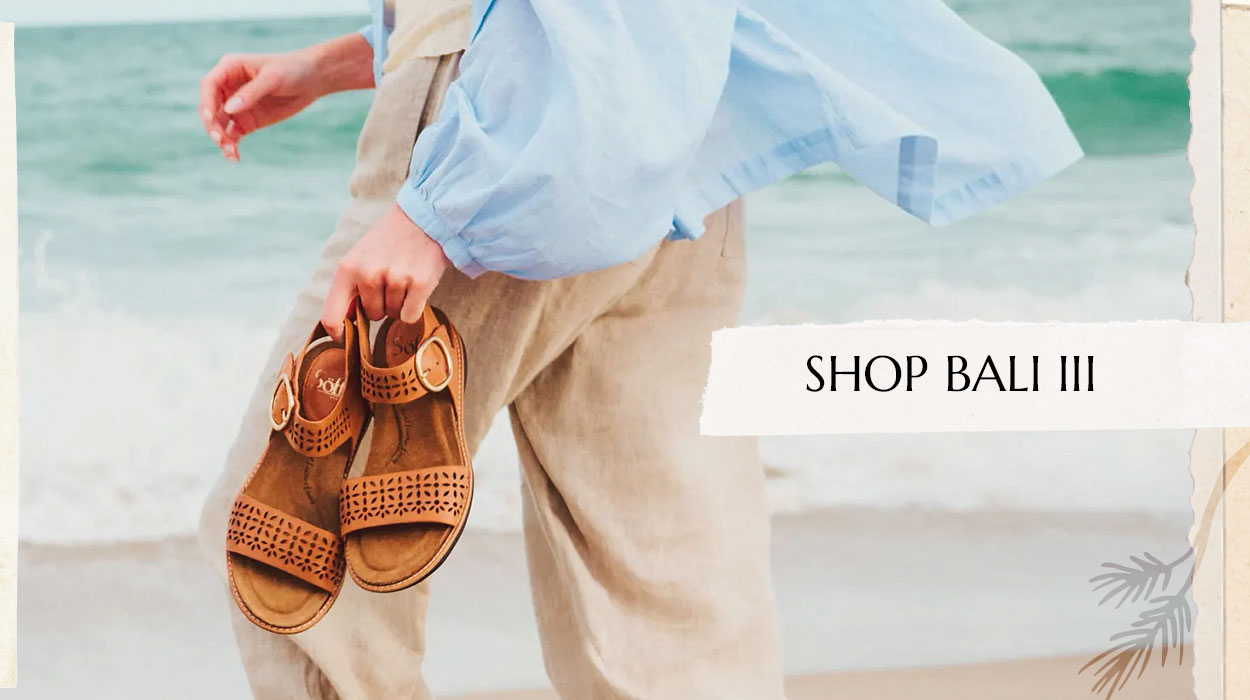 Woman wearing a blue shirt and white pants holding a pair of the Bali II sandal in brown. Shop the Bali III.